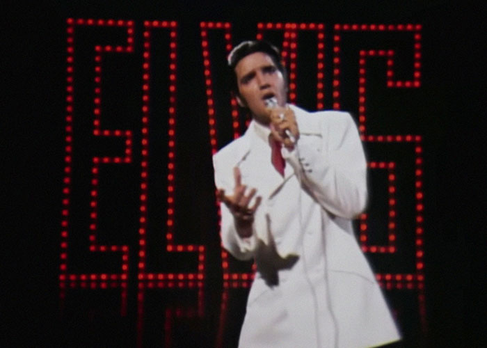 A performer in a white suit singing on stage with a large illuminated ELVIS sign in the background, capturing post-anesthesia acts.
