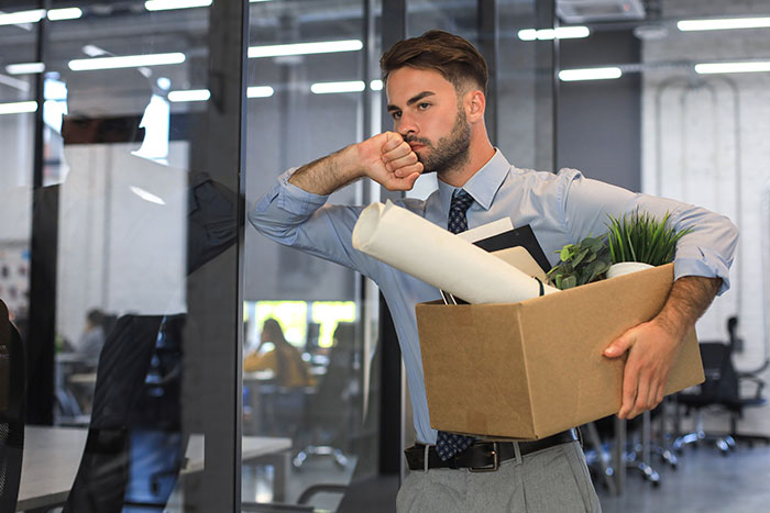 Young professional leaving office with box of belongings, showing subtle red flags indicating job insecurity. Young professional leaving office with box of belongings, showing subtle red flags indicating job insecurity.
