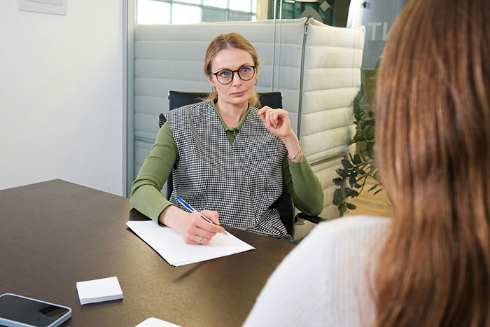 Woman in glasses conducting a serious job interview, showing subtle red flags that mean job not safe at all. Woman in glasses conducting a serious job interview, showing subtle red flags that mean job not safe at all.