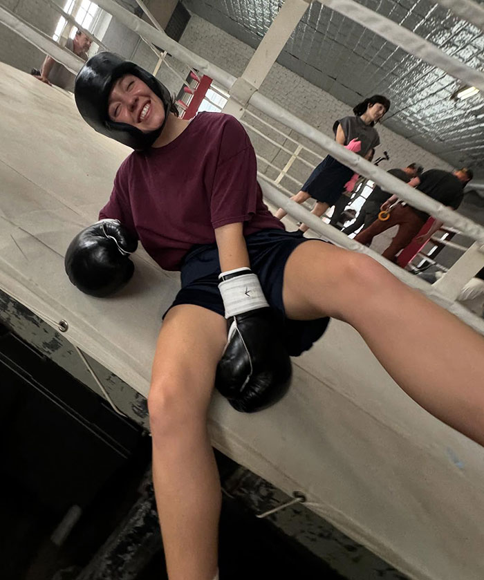 Sydney Sweeney wearing boxing gloves and helmet, smiling and relaxing in a boxing ring during a sports photoshoot.