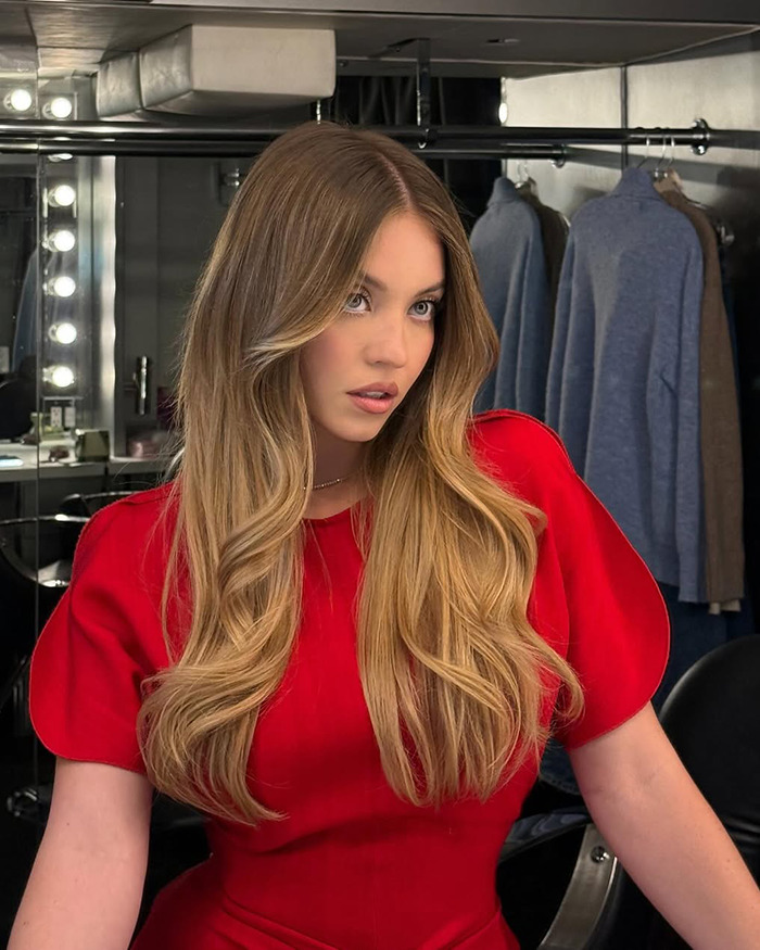 Sydney Sweeney showcasing natural beauty with styled hair and makeup in a dressing room setting with clothing in the background
