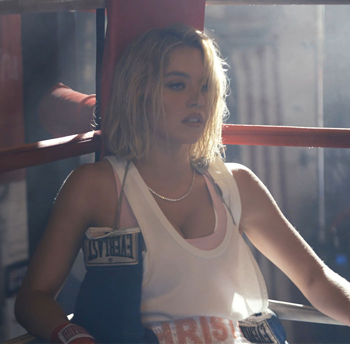 Sydney Sweeney wearing boxing gloves and sportswear, posing inside a boxing ring with moody lighting.