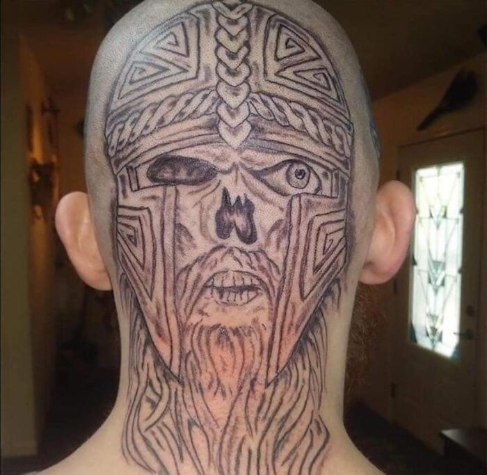 Tattoo of a poorly executed Viking helmet and face on the back of a person's shaved head, showcasing one eye closed.