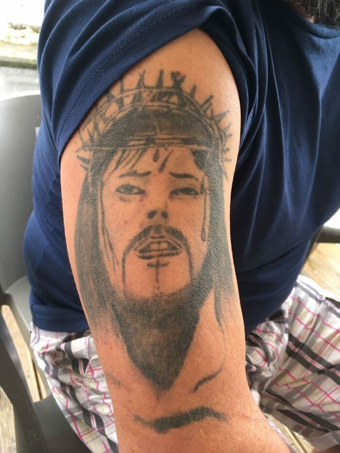 Tattoo of a poorly drawn face with a crown of thorns on a person's upper arm, representing worst tattoos collection.