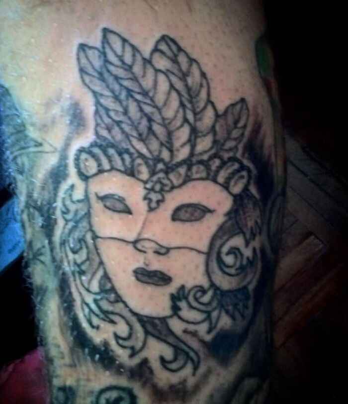 Tattoo of a mask with feathers and swirls in black ink, showcasing one of the worst tattoos in an online tattoo group.