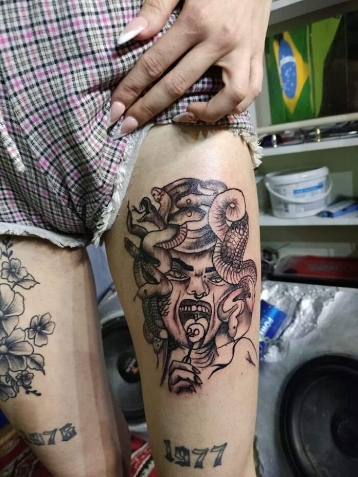 Tattoo of a Medusa-like figure with snakes and an open mouth licking a lollipop on a person's thigh.