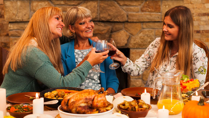 Three women clinking wine glasses at Thanksgiving dinner, with a feast including turkey and candles on the table.