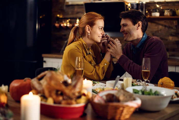 Couple holding hands at main Thanksgiving table while son looks annoyed sitting at overflow table.