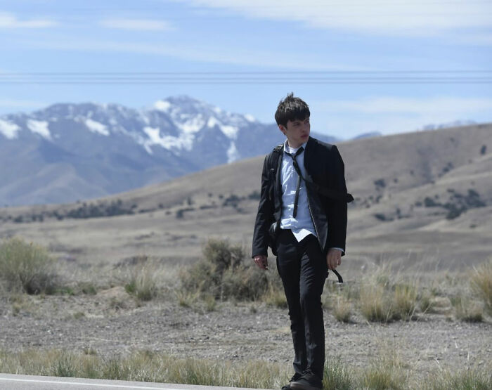 Young man in a disheveled suit walking alone on a roadside with mountains in the background, evoking a chaotic hotel room scene.