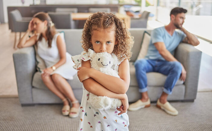 Child hugging a stuffed animal looking upset while parents sit apart, illustrating overrated and overpriced experiences.