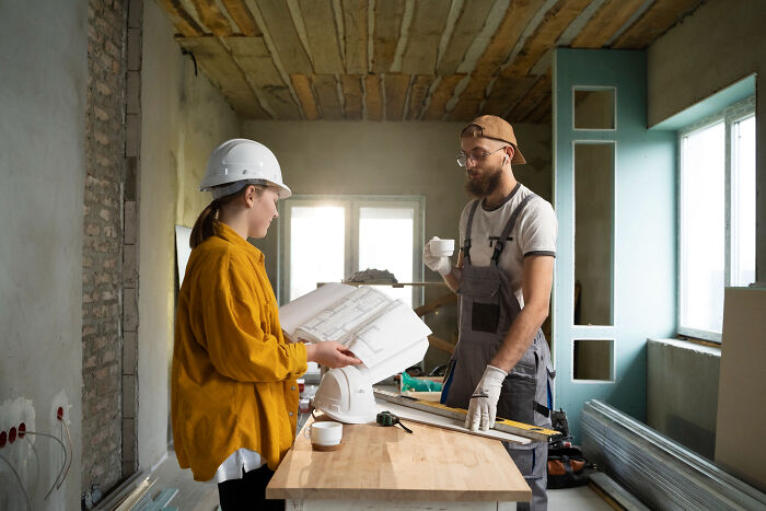 Two construction workers reviewing blueprints during renovation in an unfinished room, highlighting overrated experiences.