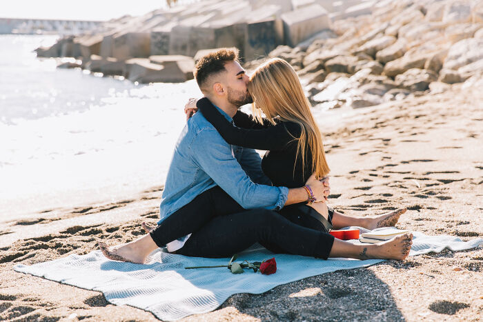 Couple sitting on beach blanket kissing with rose and book nearby, illustrating overrated must-try experiences.
