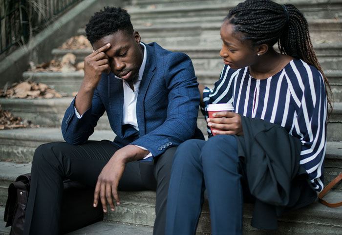 A stressed man in a blue suit sits on steps while a woman talks to him, illustrating female roasts hitting men hard.