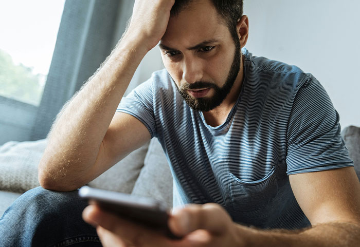 Man sitting on couch looking stressed while holding phone, reacting to female roasts that hit men hard.