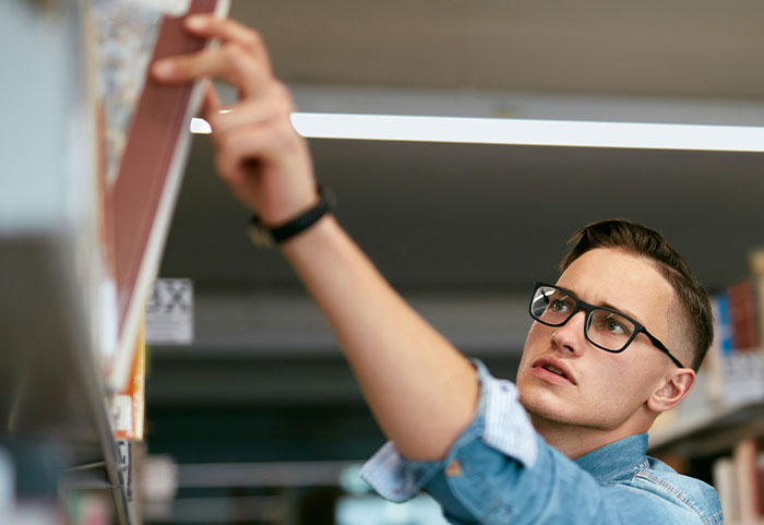 Young man wearing glasses and a denim shirt reaching for a book in a library, showcasing female roasts impact on men.