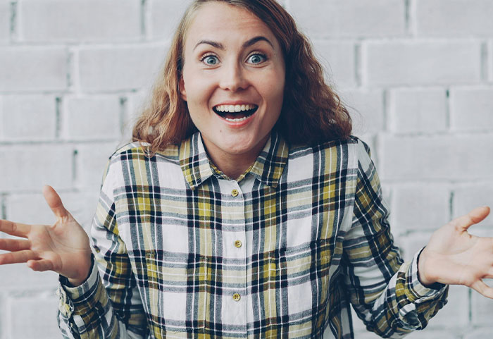 Young woman in a plaid shirt with an expressive smile and open hands, delivering female roasts with confidence.