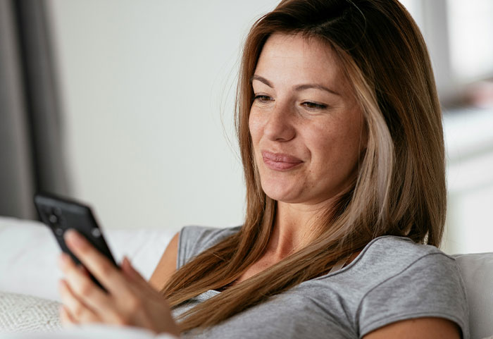 Woman in a casual grey shirt sitting on a couch, smiling subtly while reading female roasts on her phone.