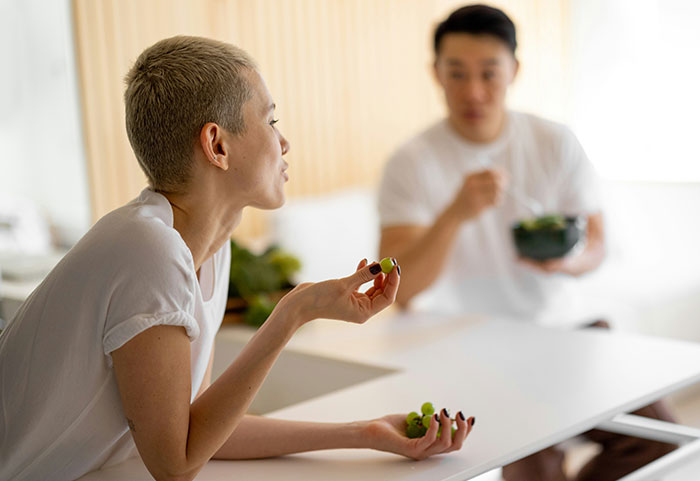 Woman with short hair eating grapes and roasting a man in casual conversation at a modern kitchen table.