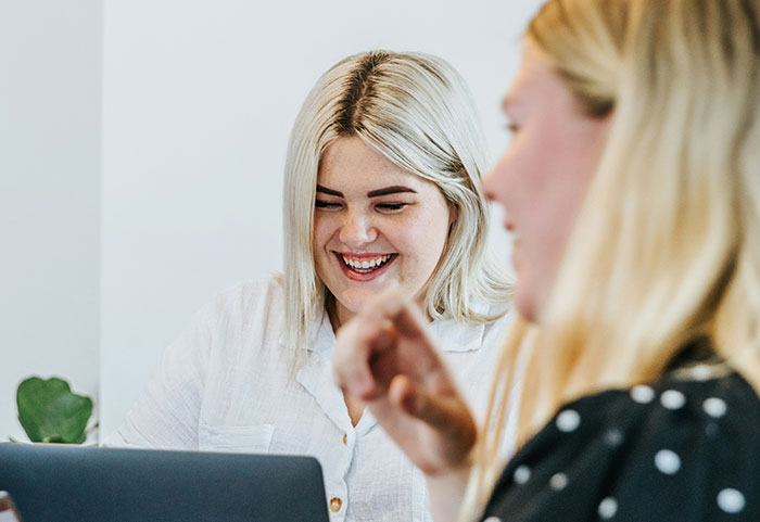 Two women laughing together while looking at a laptop, capturing female roasts hitting men hard in conversation.