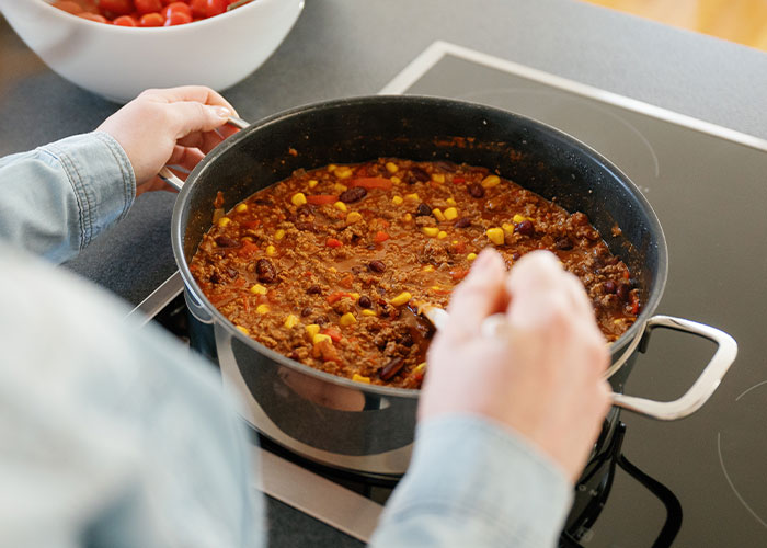 Person stirring a large pot of chili on stovetop with visible beans, corn, and ground meat ingredients.