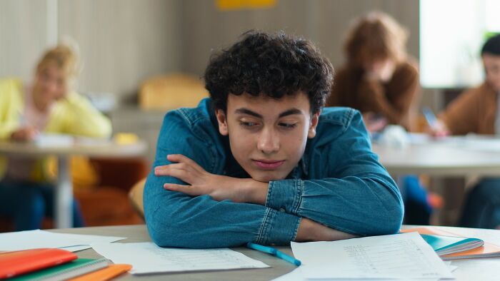 Teen boy with curly hair resting head on crossed arms, surrounded by study materials, symbolizing incredible minds without a degree.