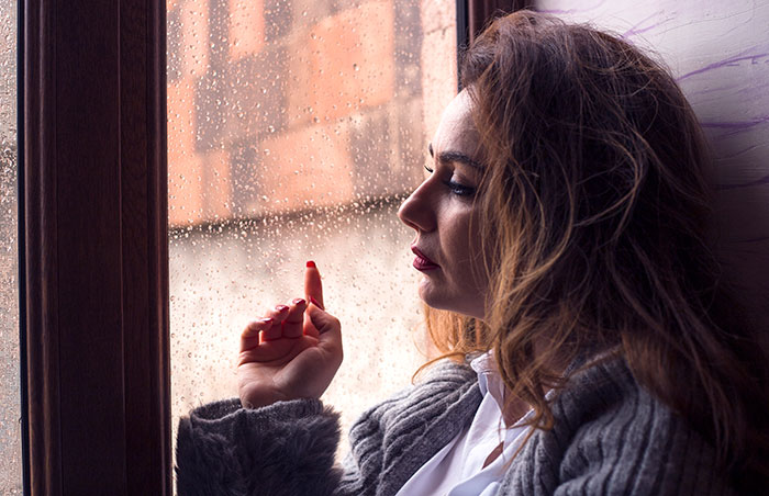 Woman in gray sweater staring sadly out a rainy window reflecting on couple&rsquo;s first free Saturday turning sour.