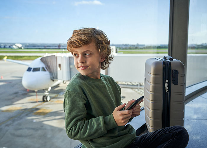 Young boy with a suitcase sitting by airport window holding phone, representing interesting today I learned facts concept.