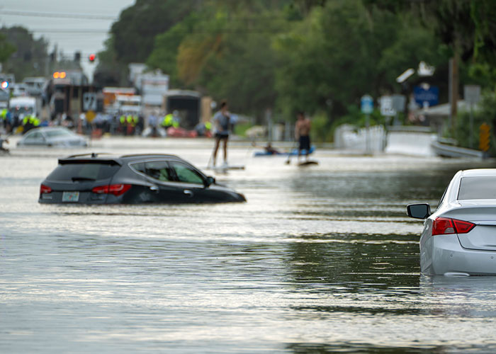 Flooded street with multiple partially submerged cars and people navigating the water in a flooded neighborhood scene.