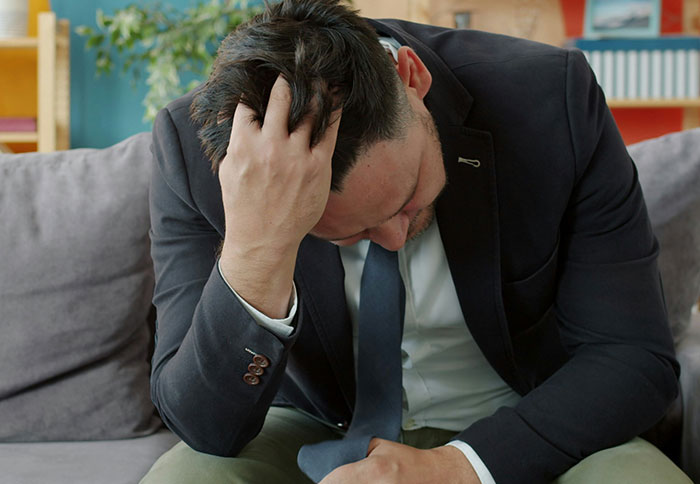 Man in business attire sitting on couch, holding head in hand, illustrating dad privileges and parental challenges.