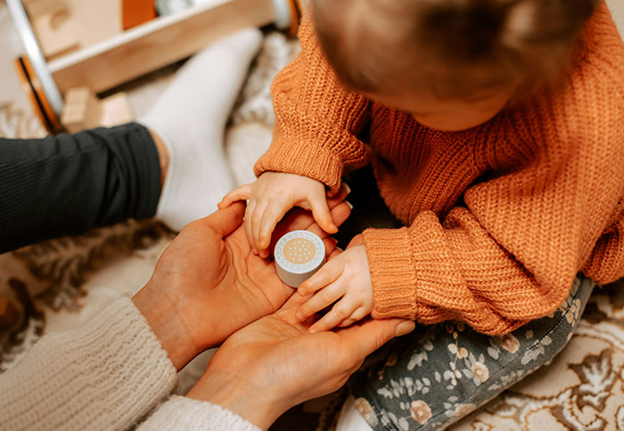 Toddler in an orange sweater holding a small container while an adult supports with both hands, highlighting dad privileges.