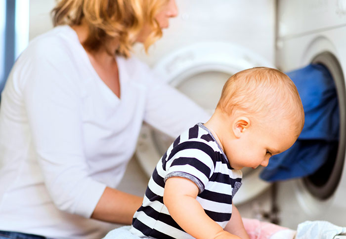 Mom multitasking near a washing machine while her toddler plays, highlighting challenges of dad privileges in parenting.