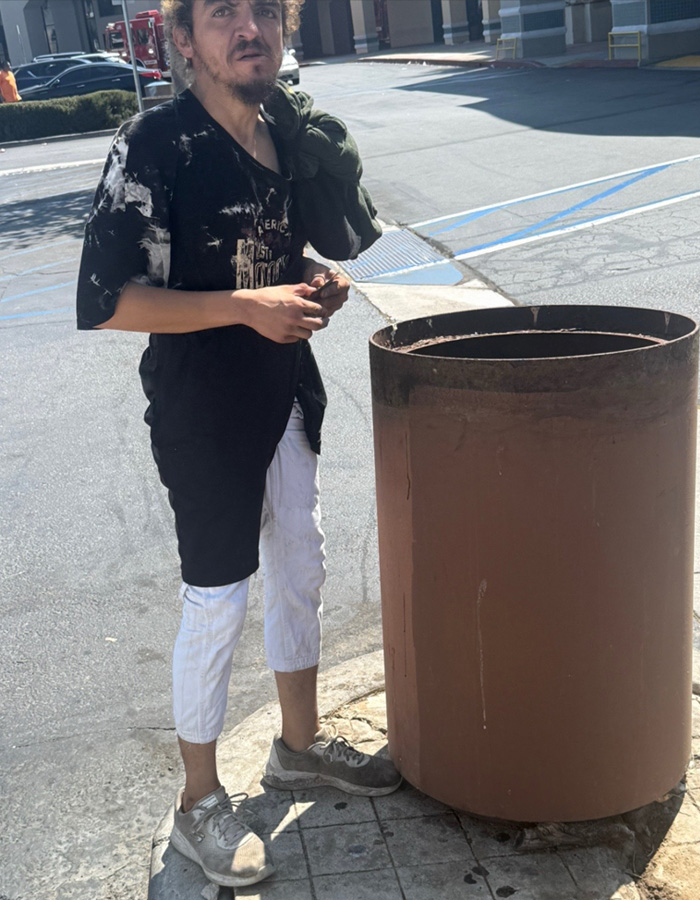 Former child actor Tylor Chase standing outdoors in casual clothes near a large rust-colored trash can on a sunny day.