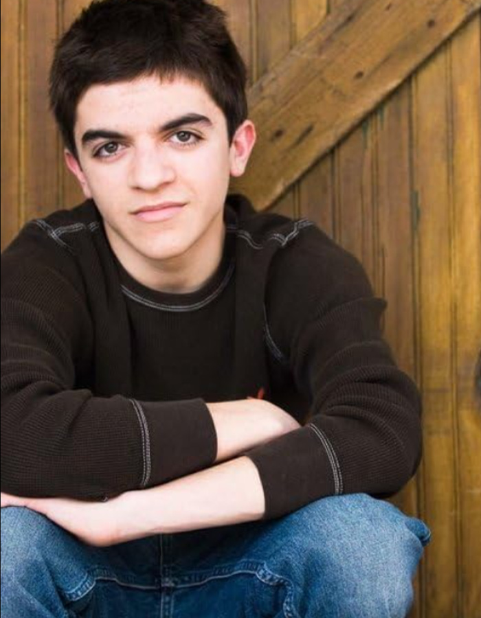 Former child actor Tylor Chase sitting against a wooden background, wearing a black sweater and blue jeans.