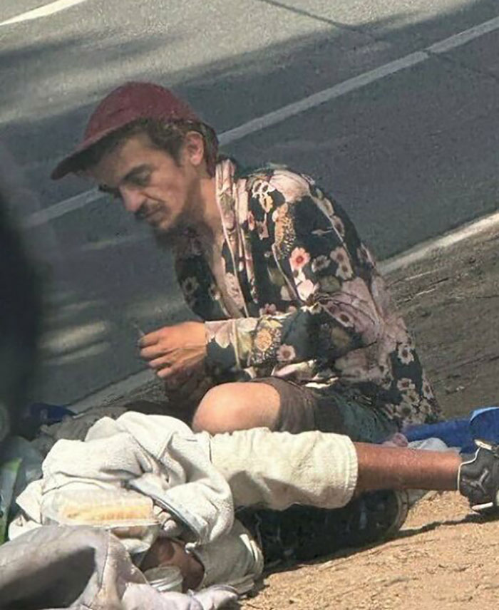 Man in floral jacket and cap sitting on street near struggling Nickelodeon star’s motel stay scene outdoors.