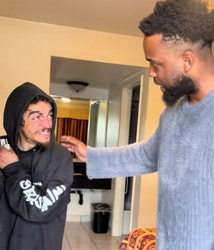 Two men in a motel room, one wearing a black hoodie and the other in a grey sweater, discussing Nickelodeon star's motel stay.