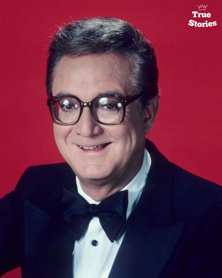 American personality wearing glasses and a black tuxedo, smiling against a bright red background in a formal portrait.