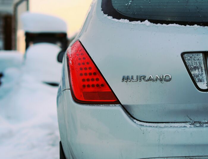 Close-up of a snow-covered gray Murano car showing rear light and badge in a winter setting, capturing strange kid's insult vibe.