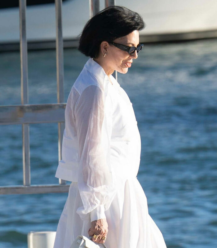 Kris Jenner in white outfit and sunglasses, walking outdoors by water, hands visible after viral $100k facelift news.