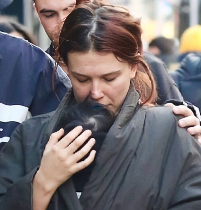 Millie Bobby Brown tenderly holding newborn daughter wrapped in dark clothing amidst a busy outdoor crowd. Millie Bobby Brown tenderly holding newborn daughter wrapped in dark clothing amidst a busy outdoor crowd.