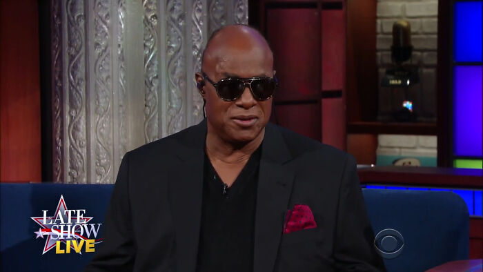 A celebrity wearing sunglasses and a black suit jacket, sharing fascinating secrets on a talk show set.