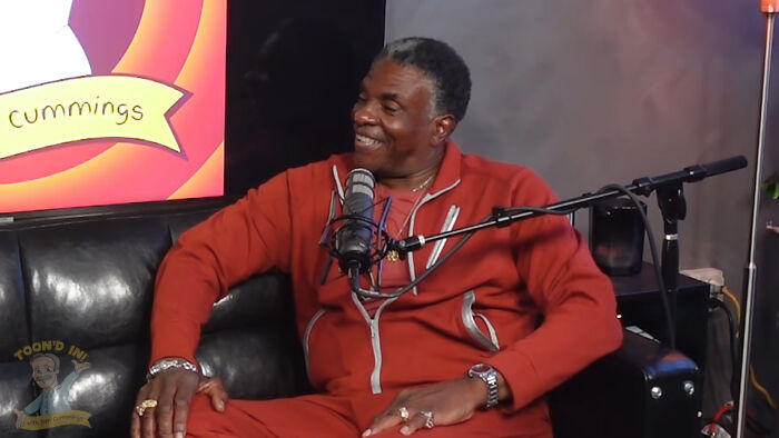 Man in red tracksuit speaking into a microphone during a podcast about fascinating secrets about celebrities.