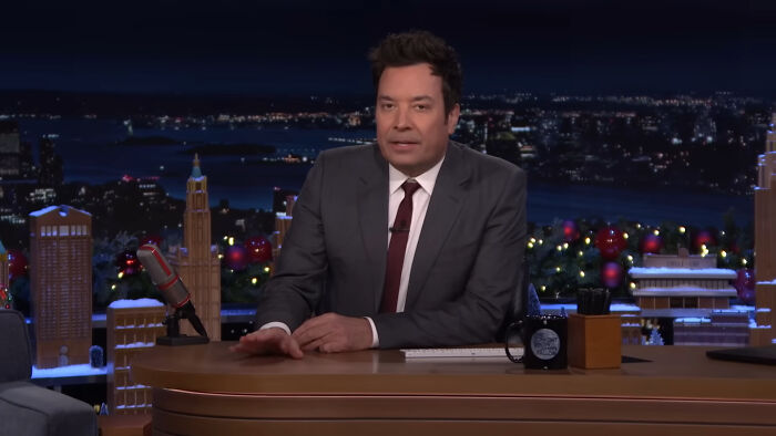 Talk show host in a dark suit discussing celebrities people think suffer on a late-night TV set.