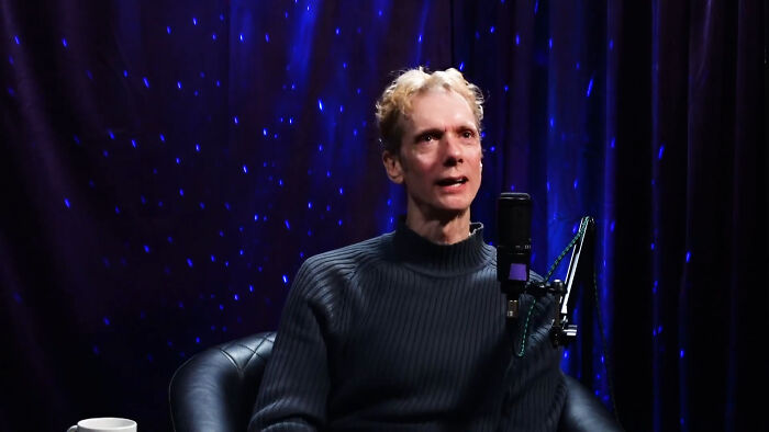 Man in a dark sweater speaking into a microphone during a recording about fascinating celebrity secrets.