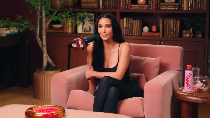 Kim Kardashian sitting on a pink armchair speaking into a microphone during a celebrities people think suffer podcast session.