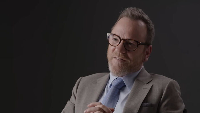Man in glasses and gray suit sharing fascinating secrets about celebrities in an interview with a dark background.