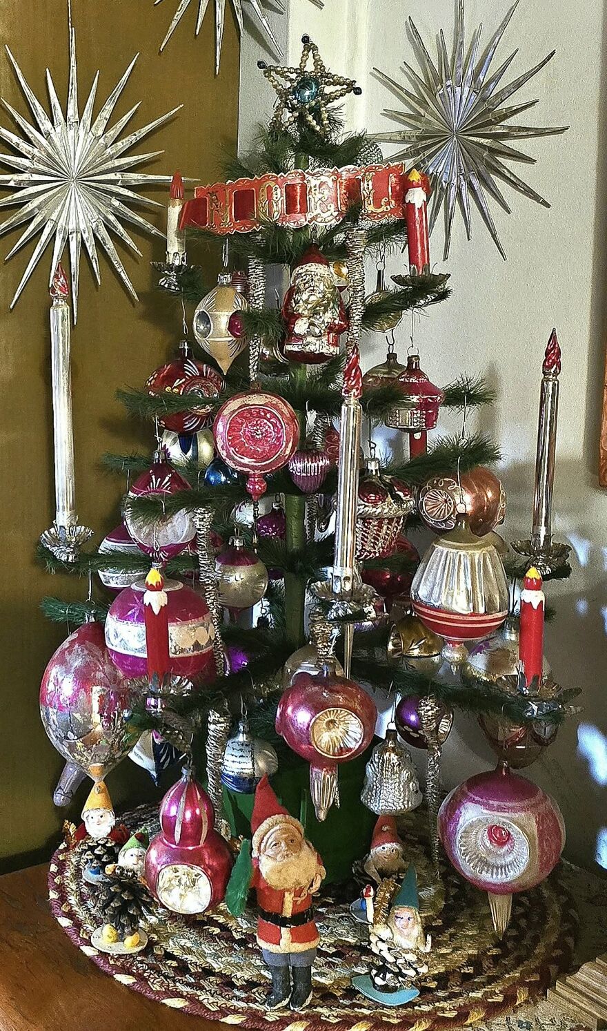 Vintage Christmas decorations on a small tree with colorful ornaments, candles, and nostalgic holiday figures on a braided rug.