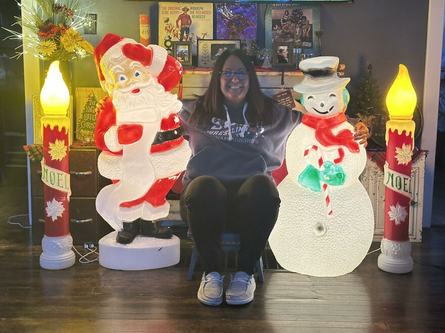 Person sitting between nostalgic vintage Christmas decorations featuring illuminated Santa, snowman, and candles indoors.