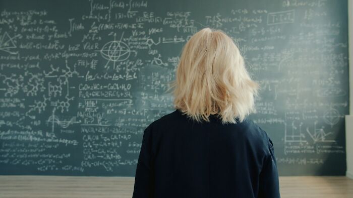 Person with blonde hair facing a chalkboard covered in complex equations representing unsolved mysteries and challenges in science.