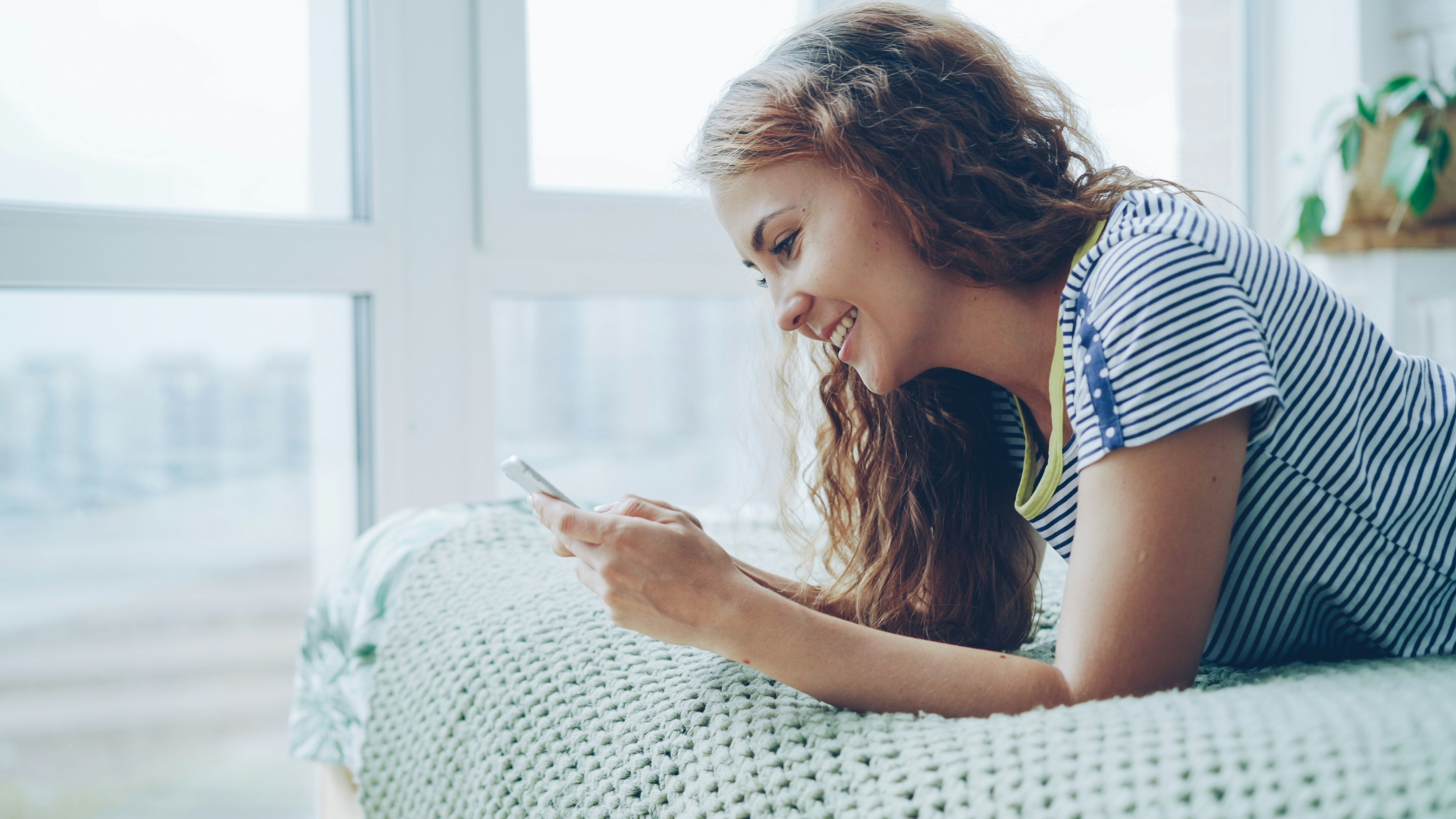 Attractive young lady is using smartphone lying on bed at home, touching screen and smiling.