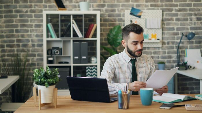 Man working at desk in office, reviewing documents with laptop and coffee mug, representing work frustrations.